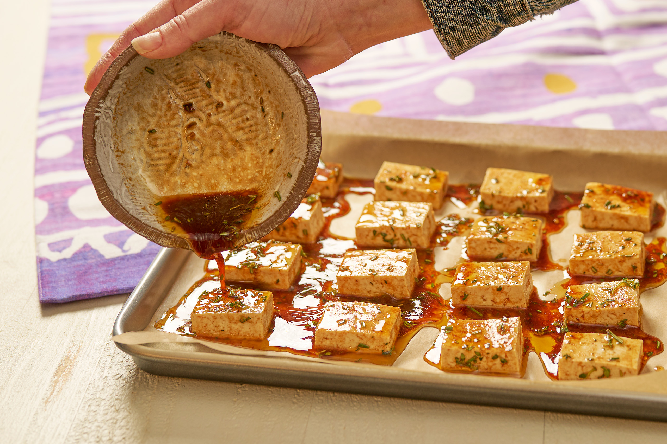 Tofu lined on a baking sheet, someone pouring marinade on top of tofu. 
