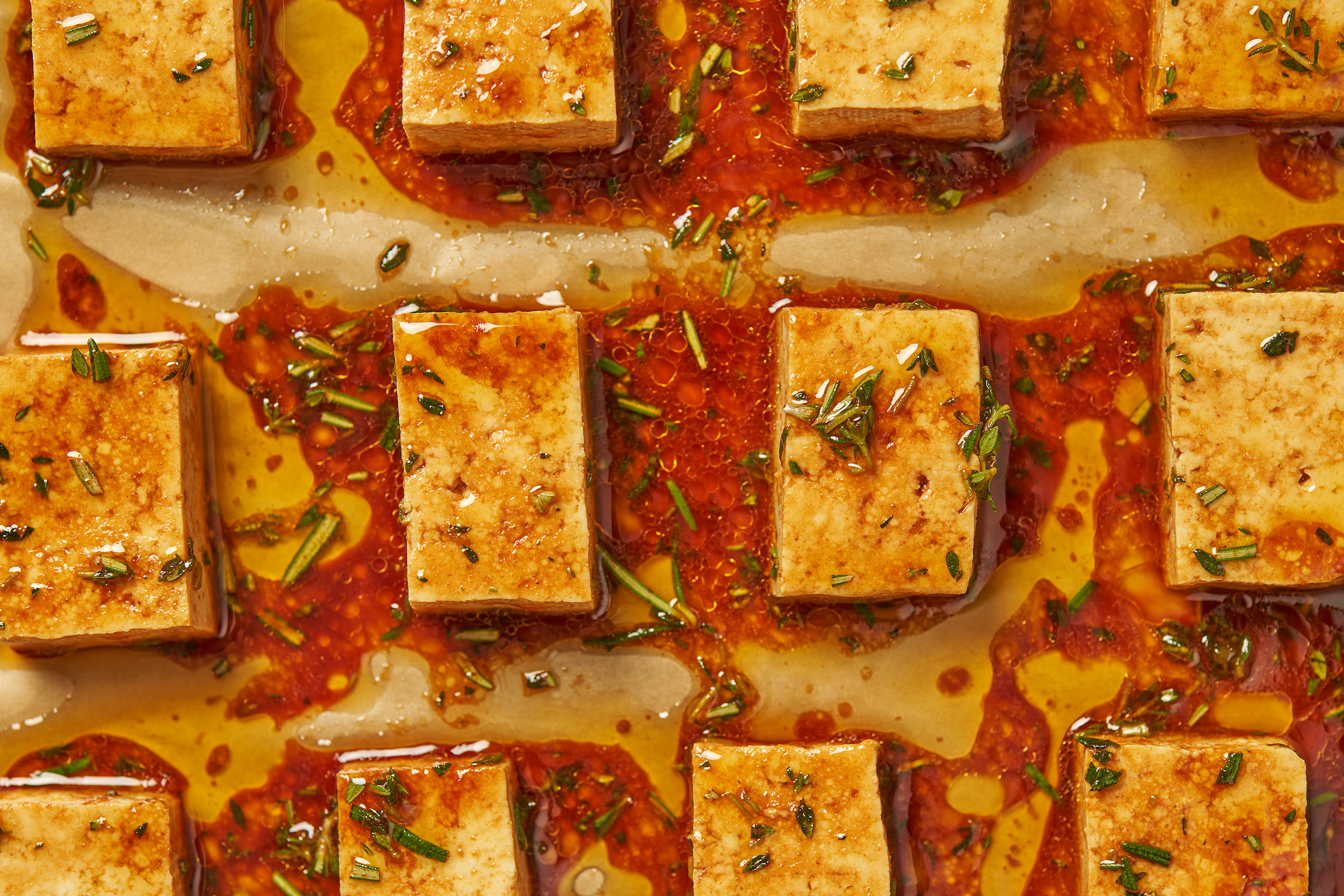 Tofu slabs arranged on a parchment-lined baking sheet with a red, herby marinade. 